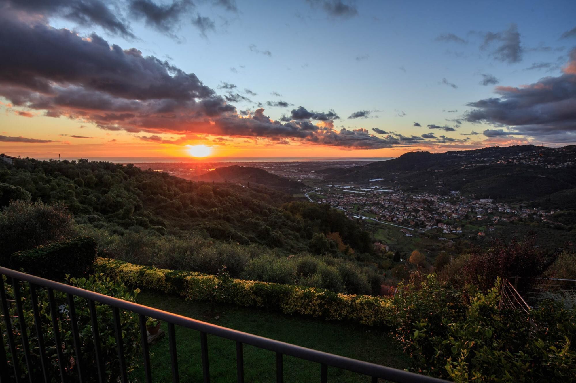 Vista panoramica di una città al tramonto, con in primo piano dolci colline verdi e una ringhiera.