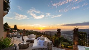 Une terrasse avec un canapé en osier, une table à manger et une vue sur le coucher de soleil au-dessus de la ville.