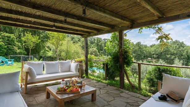 Uma área de estar coberta ao ar livre, com um banco de madeira, um sofá branco e uma mesa de pedra com frutas e bebidas.
