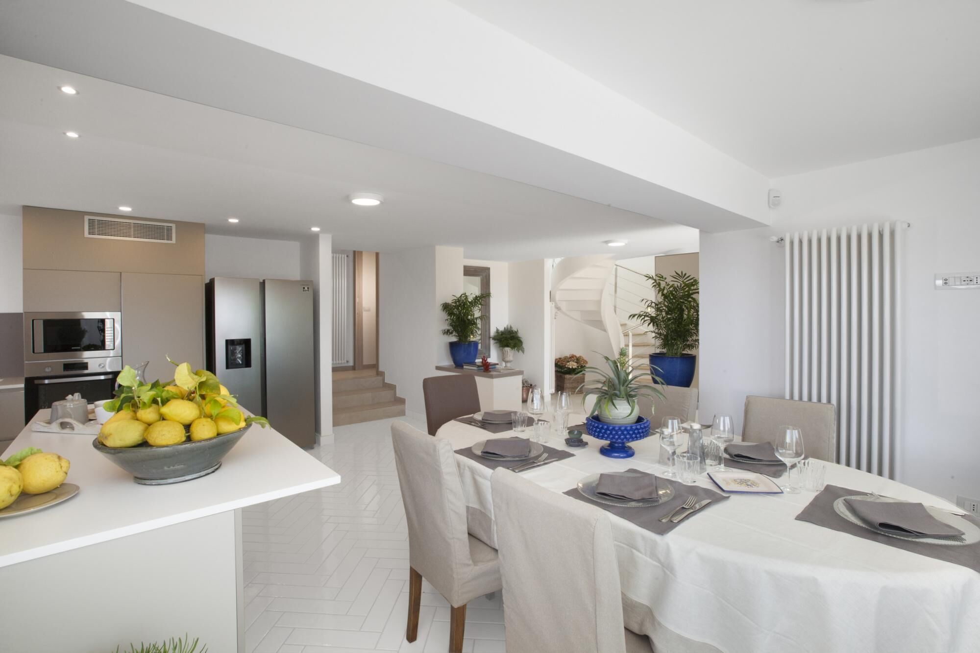 A modern dining area with a white table set for four, a kitchen with a microwave and a fruit bowl, and a staircase leading to an upper level.