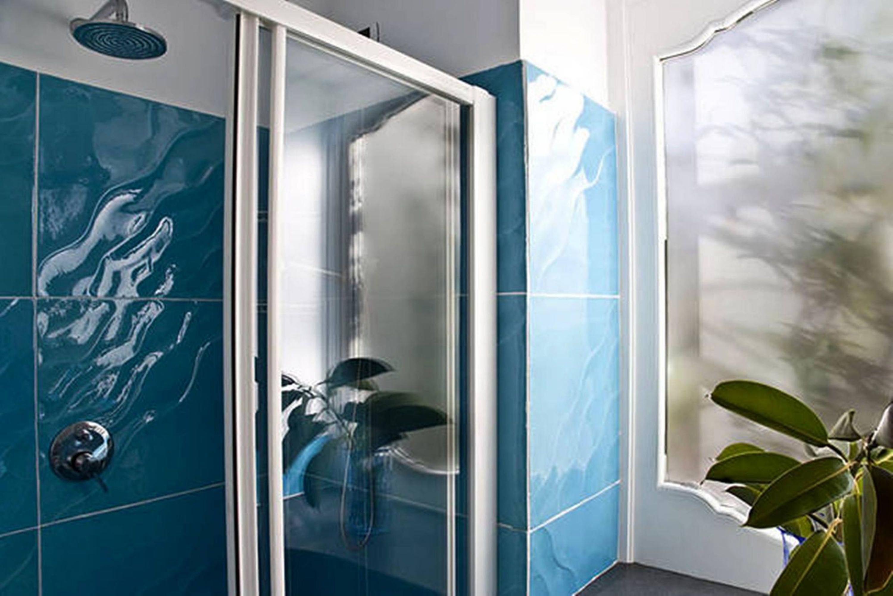 A bathroom with a glass-enclosed shower, blue tiles, and a plant near the window.