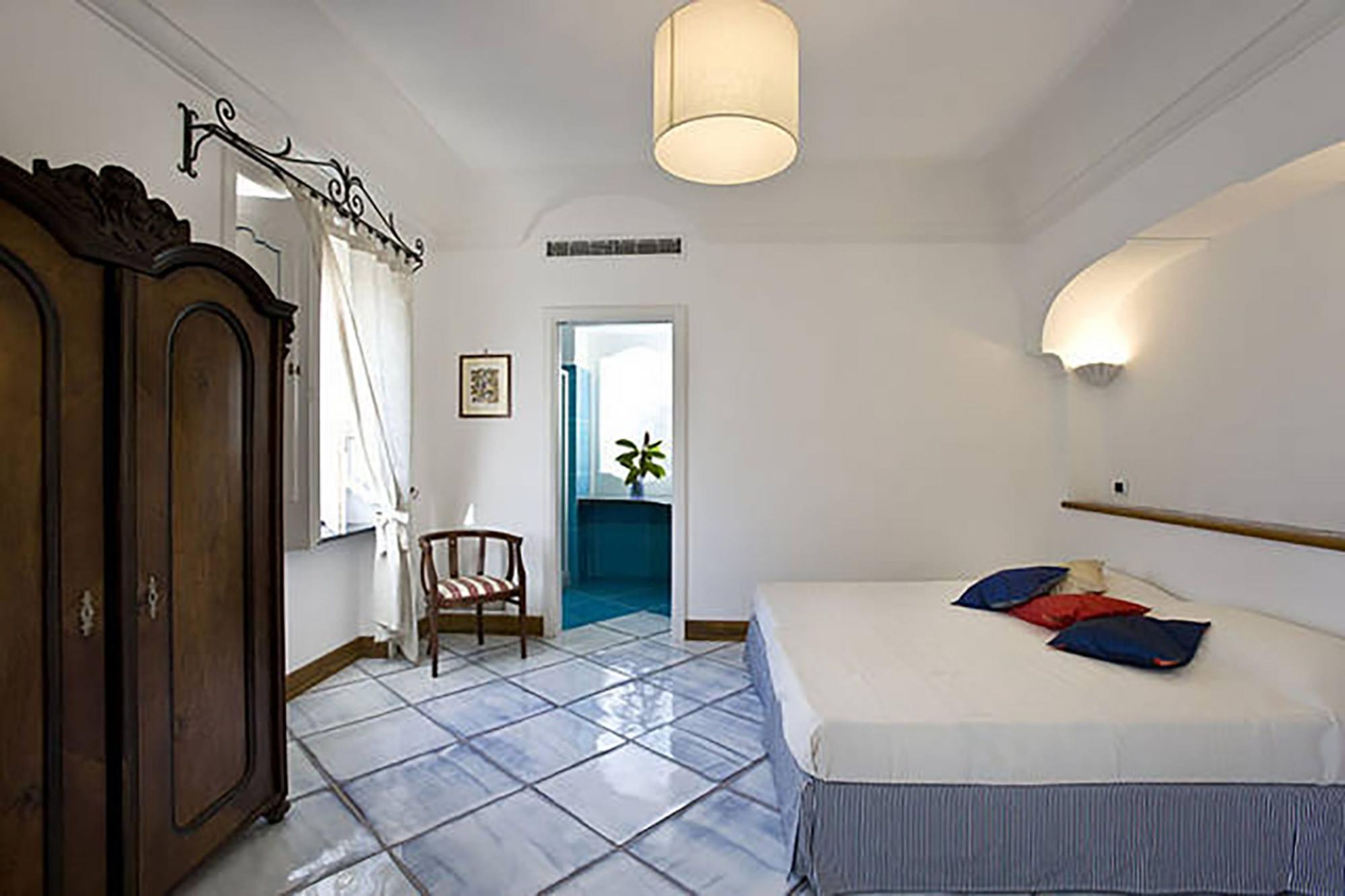 A bedroom with a bed, a wardrobe, a chair, a door leading to a bathroom, and a hanging light fixture.
