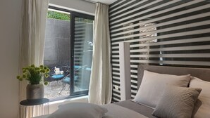 A bedroom with a bed, a window with curtains, a potted plant, and a view of an outdoor area with chairs.