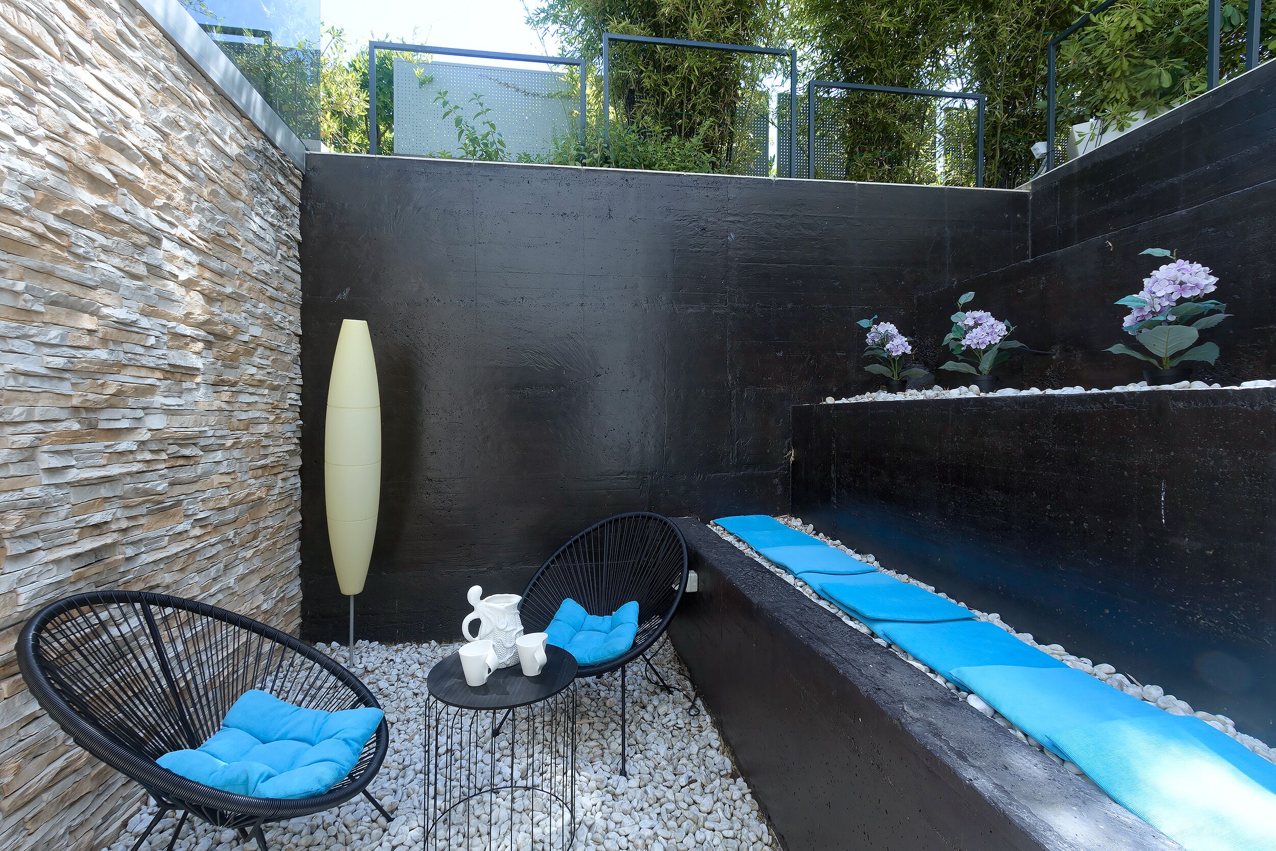 Un espace de détente extérieur moderne, avec un mur en pierre, un fauteuil noir rembourré, un fauteuil bleu rembourré et une petite table sur laquelle se trouvent des tasses blanches et une carafe.