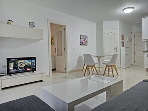 A modern living room with a flat-screen TV, a glass-top coffee table, and a dining area with two chairs.