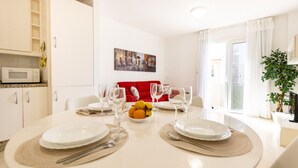 A dining area with a round table set for four, white plates, cutlery, and glasses. A bowl of fruit is on the table, and a red sofa is in the background. - TIJERITAS COOL SEA BREEZE (Los Cristianos)
