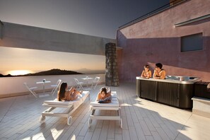 A rooftop terrace with lounge chairs, a bar area, and a view of the sunset over a hilly landscape.