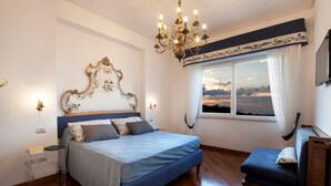 Een slaapkamer met een bed, nachtkastje, lamp en uitzicht op een zonsondergang door een raam.