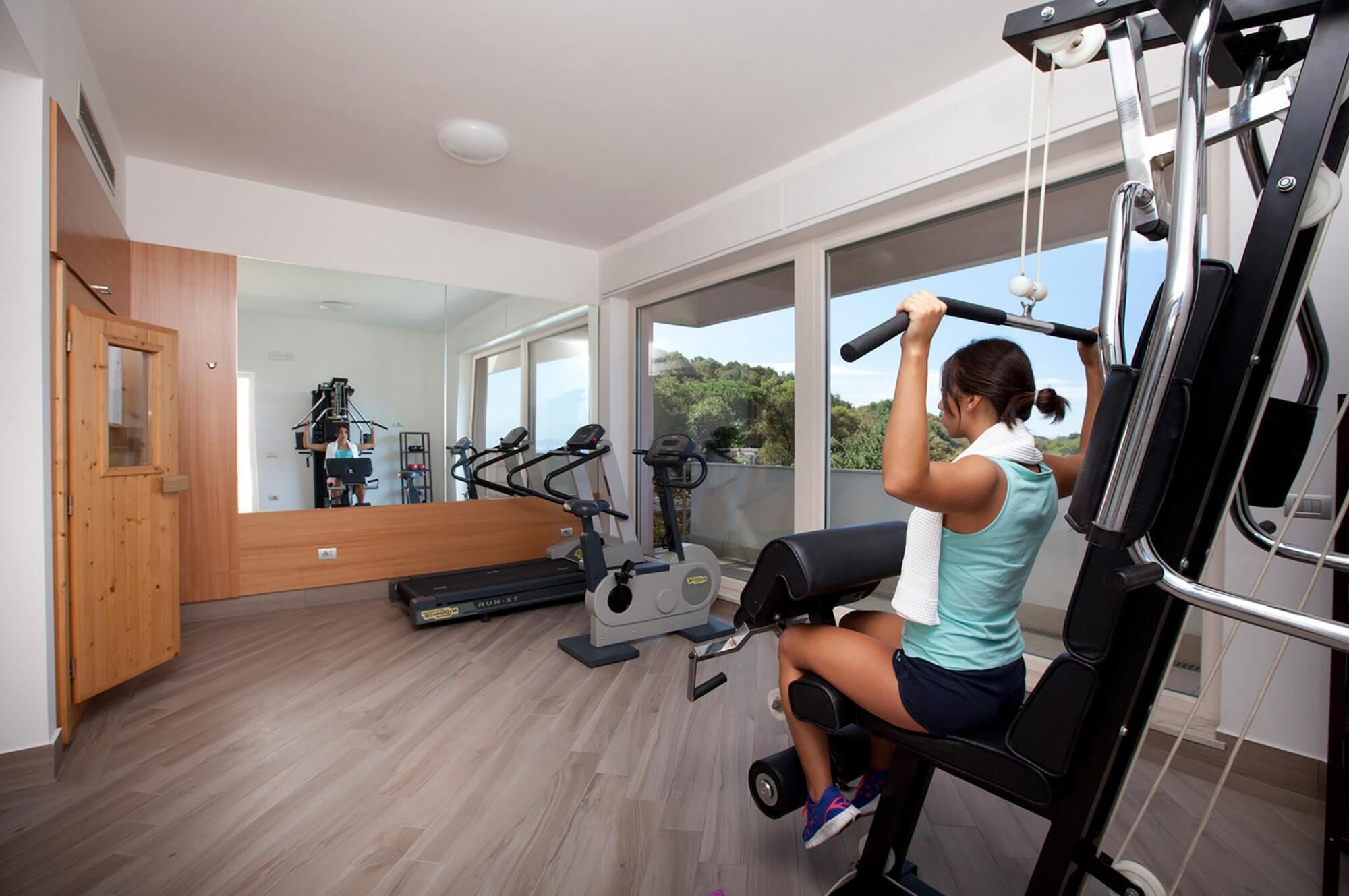 A gym with exercise equipment, a treadmill, and a large window with a view of trees.