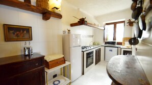 A compact kitchen with a refrigerator, stove, oven, and sink. There is a wooden shelf above the sink and a wooden table in the foreground. - Villa Posidonia in Massa Lubrense (Massa Lubrense)
