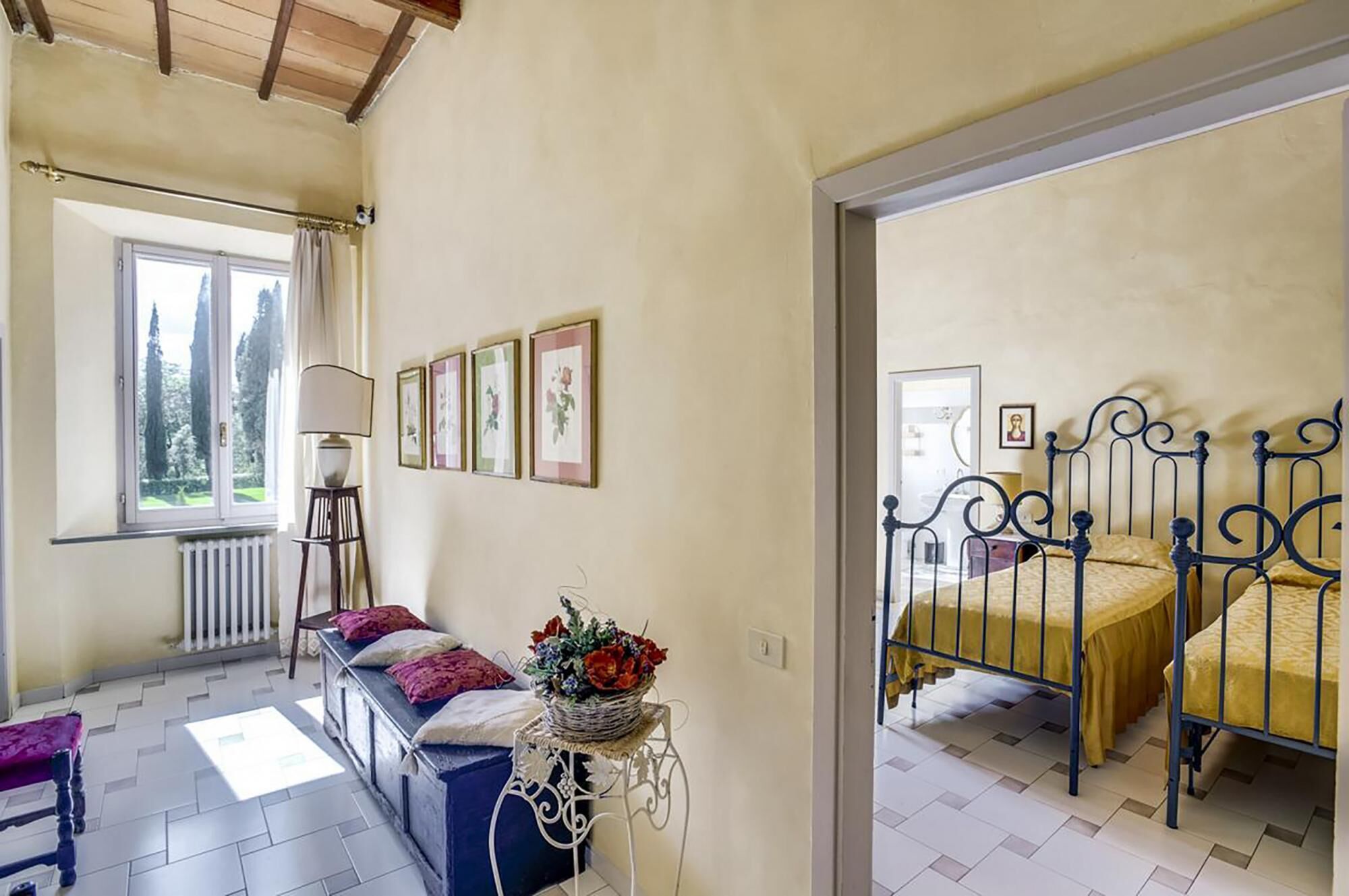 Een slaapkamer met een bed, een bankje, een tafel met een mand vol bloemen en een raam met uitzicht op bomen.