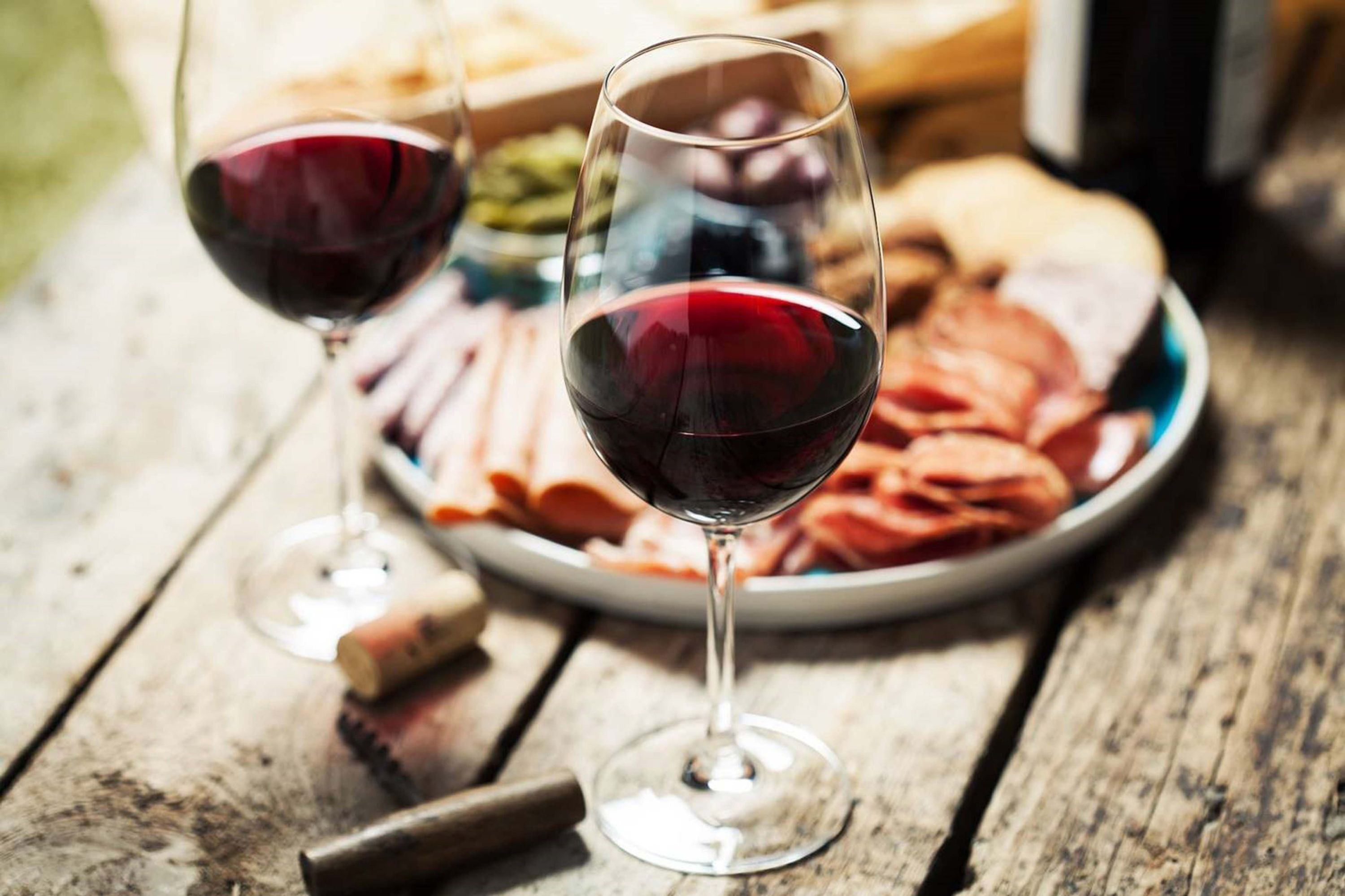 Dos copas de vino sobre una mesa de madera, con un plato de embutidos y una botella al fondo.