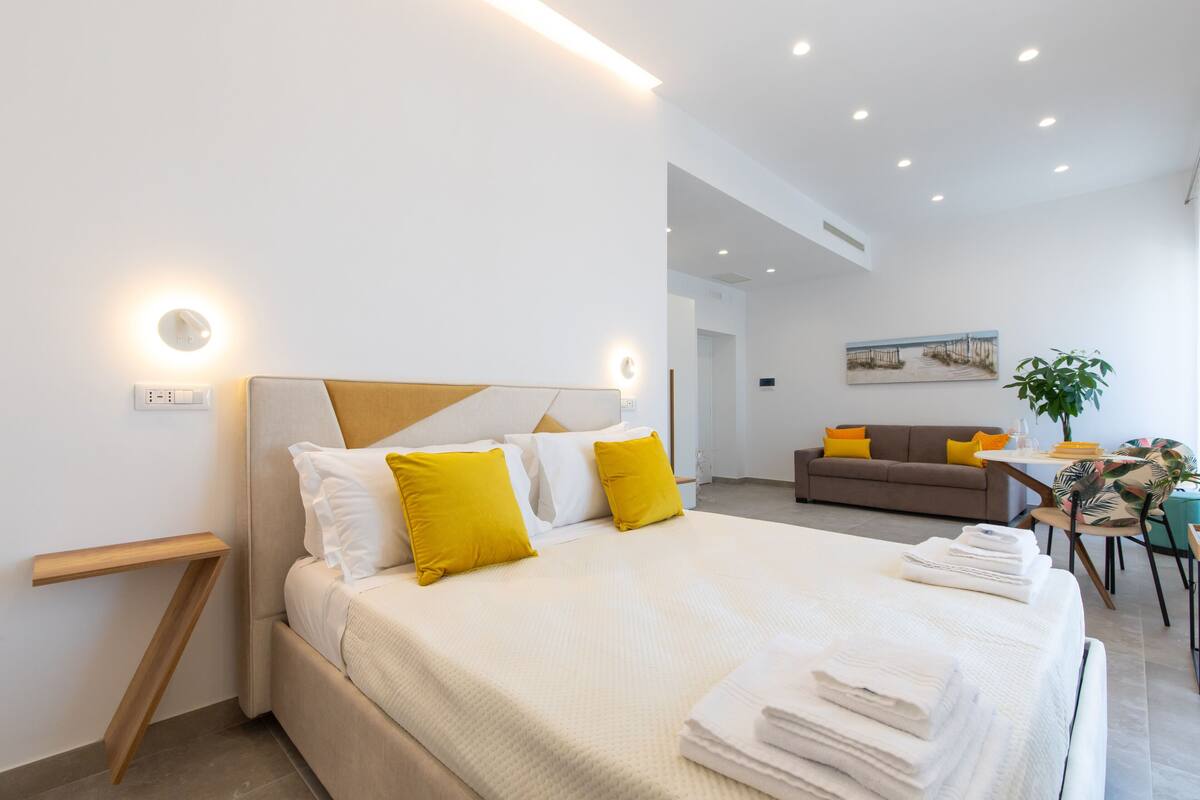 A modern hotel room with a large bed, a bedside table, a sofa, and a dining area.