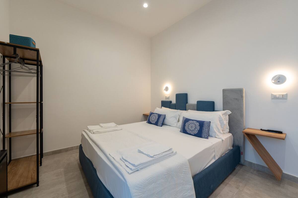 A neatly arranged bedroom with a bed, a bedside table, a wall-mounted shelf, and a wardrobe.