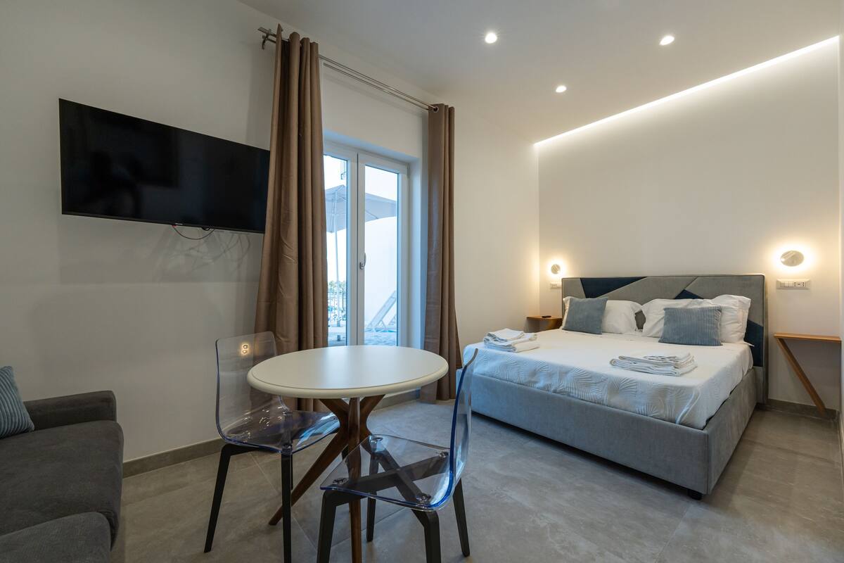 A modern hotel room with a bed, a TV mounted on the wall, a dining table with chairs, and a window with curtains.