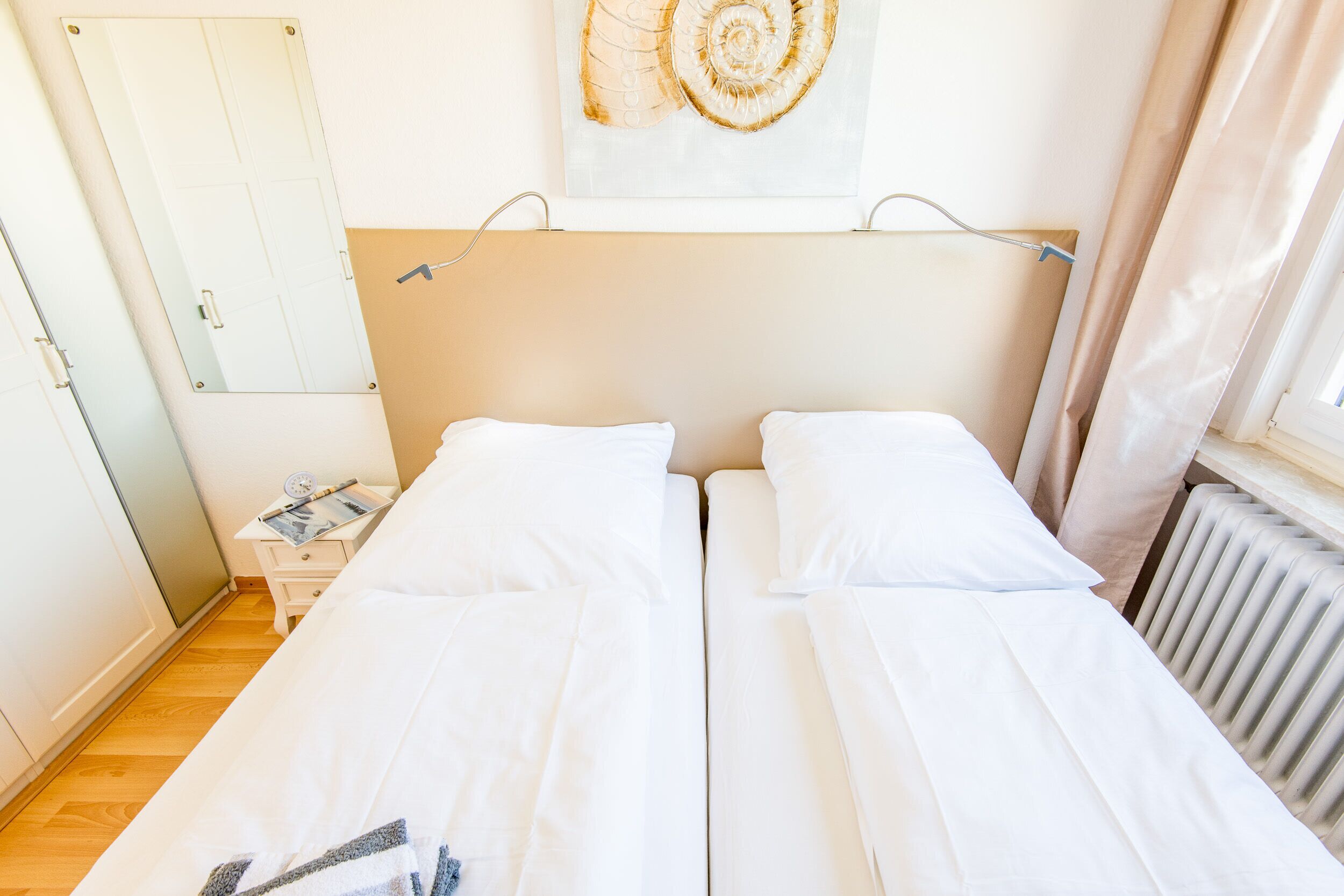 Een tweepersoonsbed met witte lakens, een nachtkastje met een lamp, een aan de muur hangend kunstwerk en een radiator.