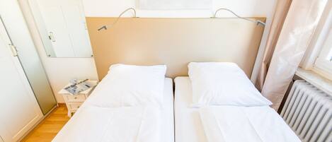 Een tweepersoonsbed met witte lakens, een nachtkastje met een lamp, een aan de muur hangend kunstwerk en een radiator.