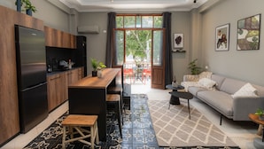 Eine moderne Küche mit integriertem Kühlschrank, Holzschränken und einer zentralen Kochinsel mit Barhocker. Es gibt einen Essbereich mit Tisch und Stühlen, eine Couch und einen kleinen Beistelltisch. Der Raum hat große Fenster mit Vorhängen, und der Boden ist mit einem geometrischen Muster gefliest.