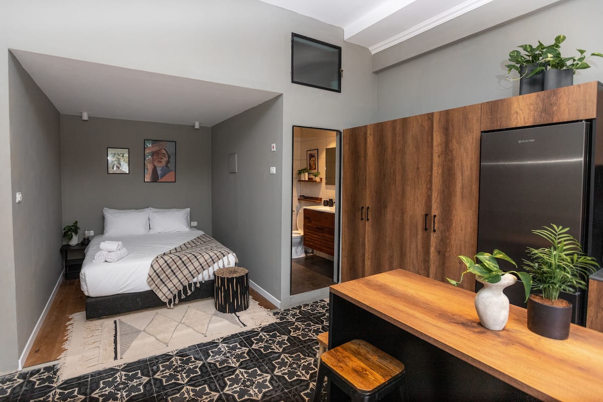 A compact bedroom with a bed, bedside table, and a small stool. A wooden wardrobe and a black refrigerator are also visible.