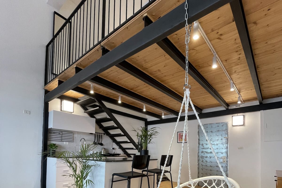 A spacious loft with a high ceiling, exposed wooden beams, a kitchen area, and a hanging macrame chair.