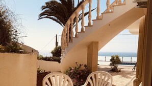 A covered outdoor area with a spiral staircase, white plastic chairs, and a view of the sea. - Mae Home-Galaxy (Foz do Arelho)
