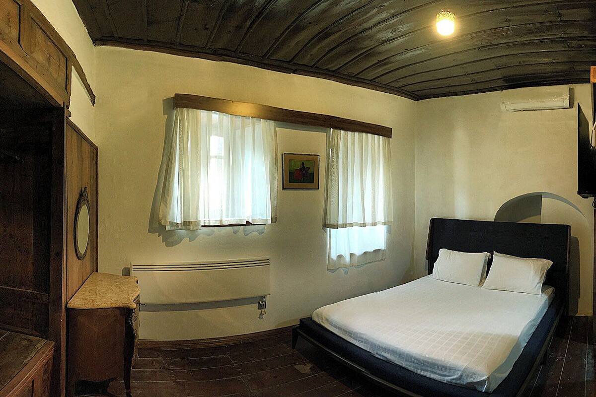 A bedroom with a bed, a wooden wardrobe, a radiator, and two windows with curtains.
