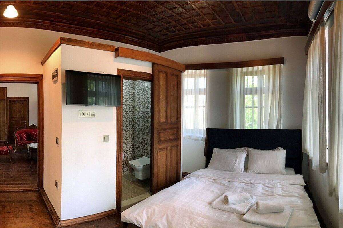 A bedroom with a large bed, wooden headboard, a TV mounted on the wall, and a bathroom visible through an open door.