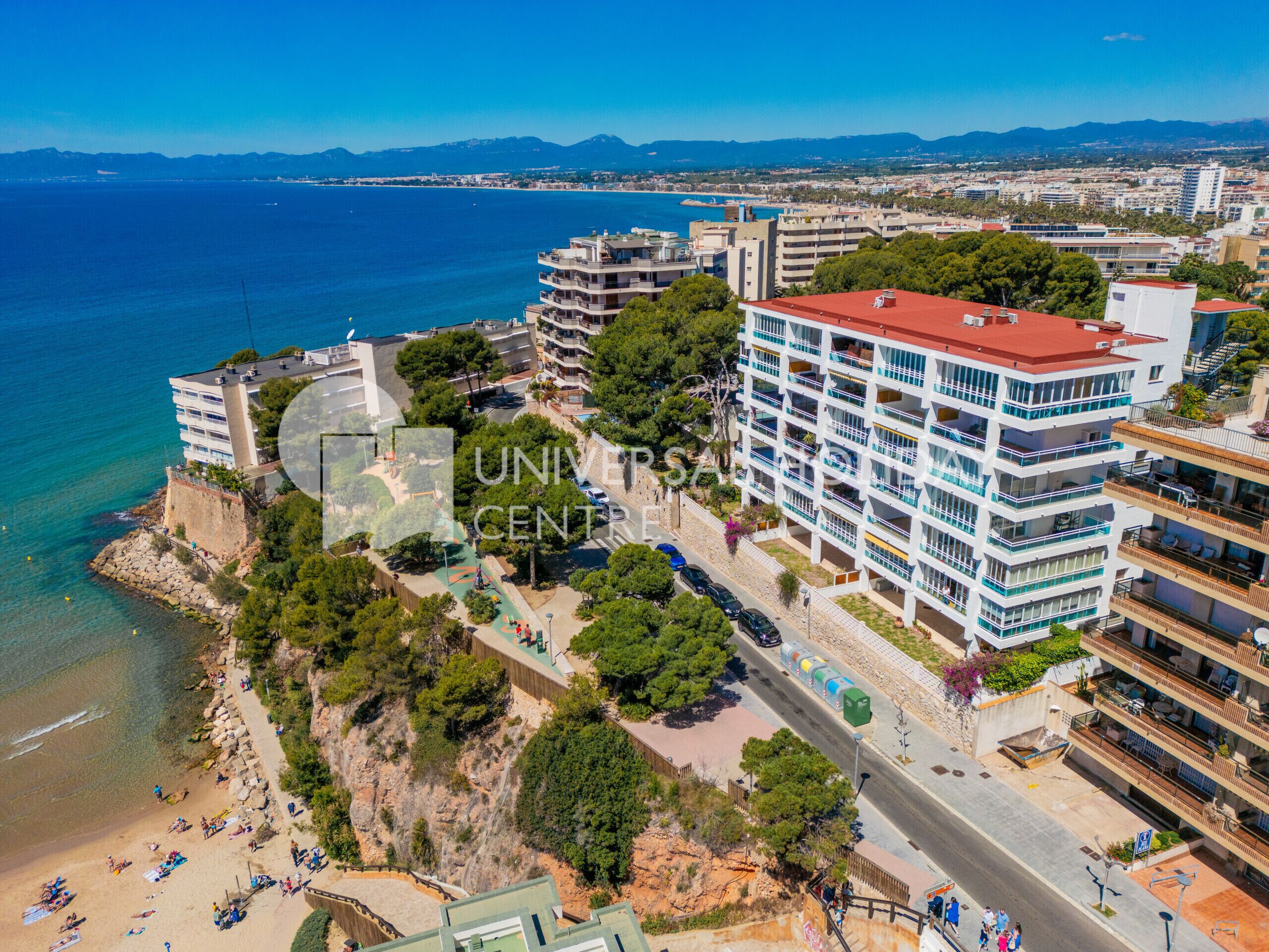 Un paisaje urbano costero con un hotel de varios pisos destacado, que cuenta con balcones, una playa y una vista despejada del mar.