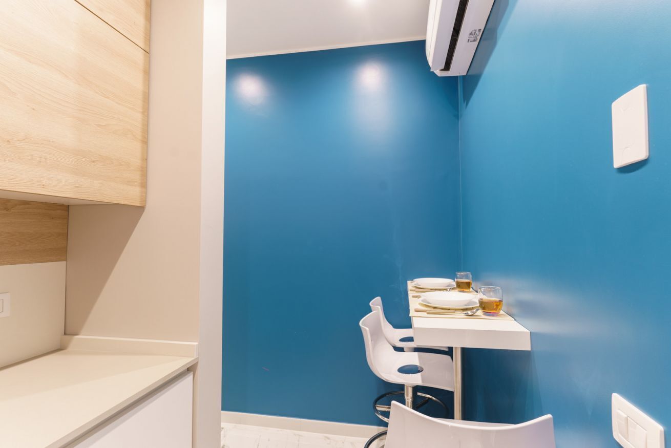 A small room with a blue wall, a white table with a chair, and a white air conditioning unit.