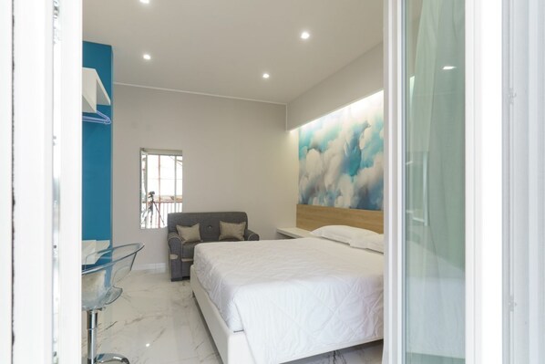 A modern bedroom with a large bed, a grey sofa, a glass table, and a wall with a cloud painting.