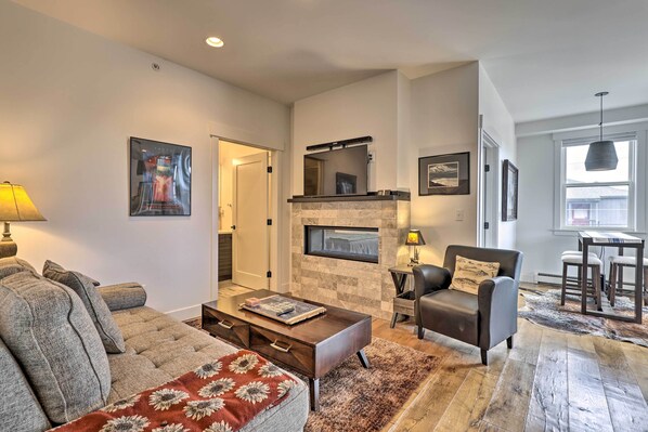 A modern living room with a sofa, coffee table, fireplace, and dining area. - Grand Lake Condo w/ Balcony & Mountain Views (Grand Lake)