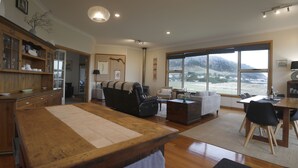 A spacious living room with a wooden dining table, a sofa, and a view of a mountainous landscape through large windows. - Stanley Beach House with Stunning Views of the Nut! (Stanley)