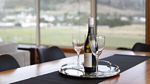 A table with a wine bottle and two glasses on a tray, set against a backdrop of a scenic view through large windows. - Stanley Beach House with Stunning Views of the Nut! (Stanley)