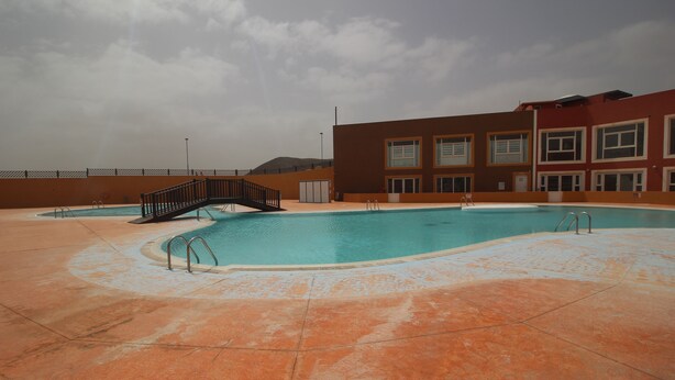 A swimming pool with a wooden bridge, surrounded by a building with multiple windows and a tiled floor. - Ideal apartment for large families and groups of friends (Corralejo)