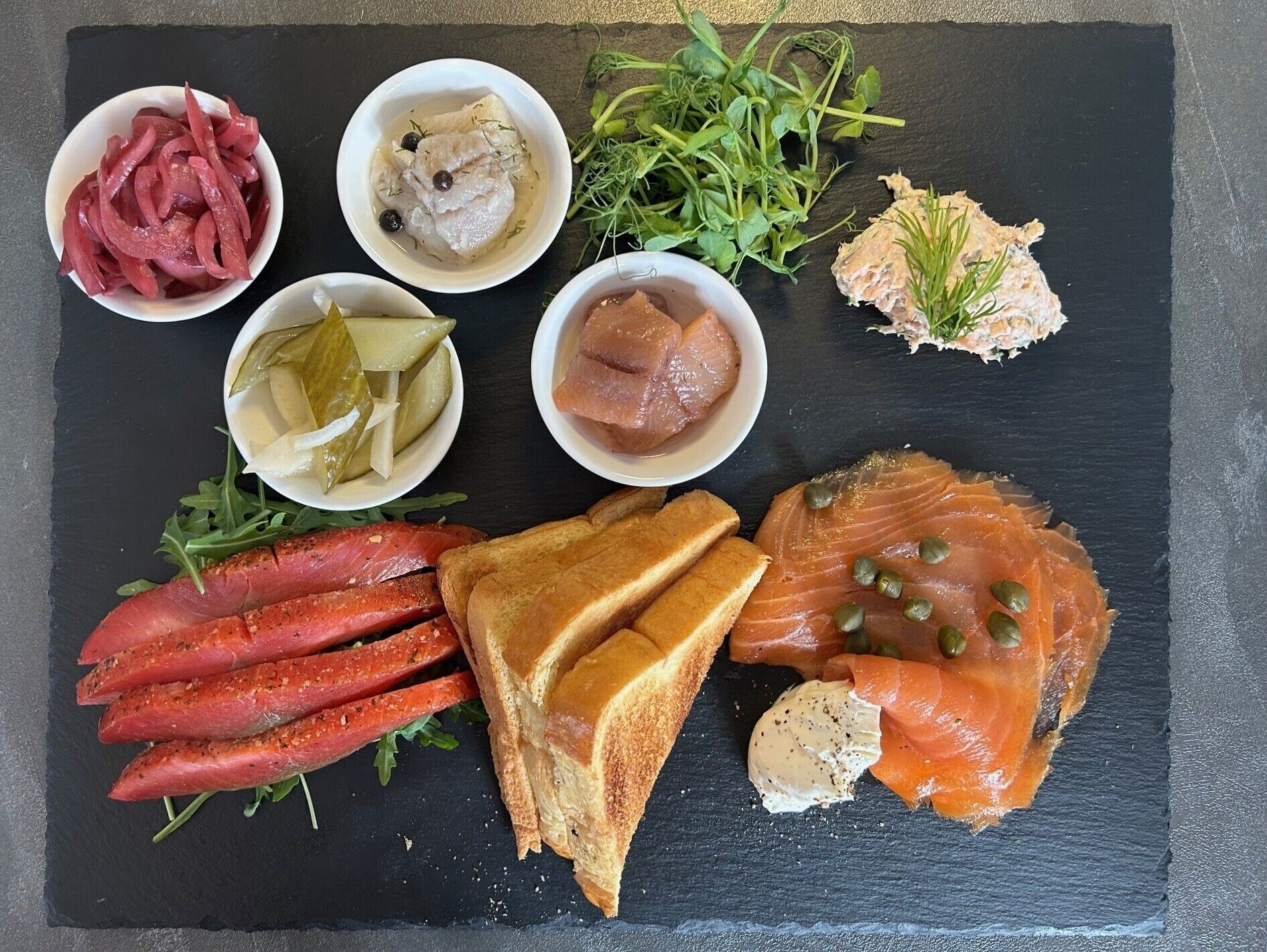 A variety of food items including smoked salmon, pickles, and bread on a slate board.