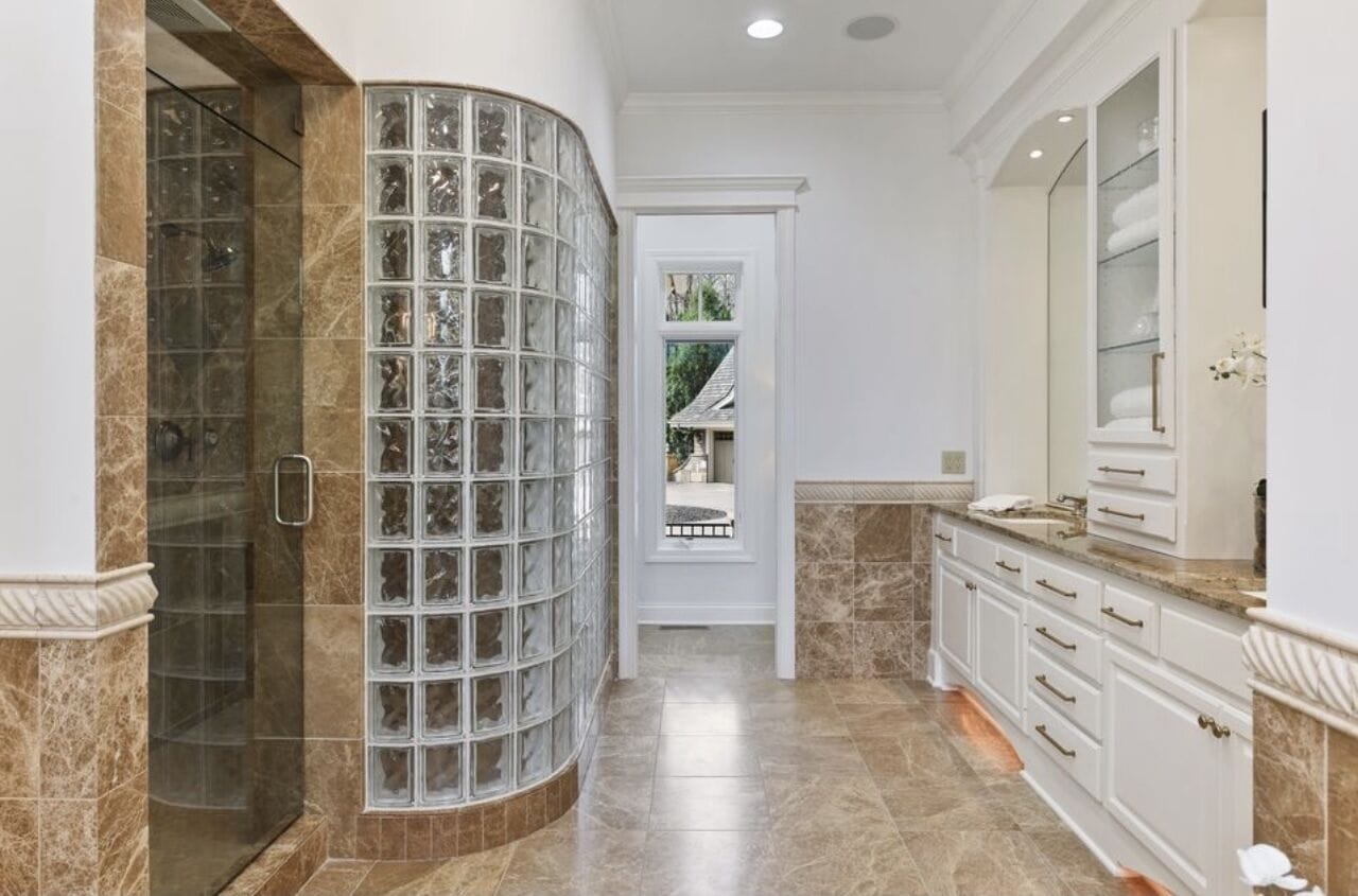 A bathroom with a glass block shower enclosure, marble flooring, and white cabinetry.