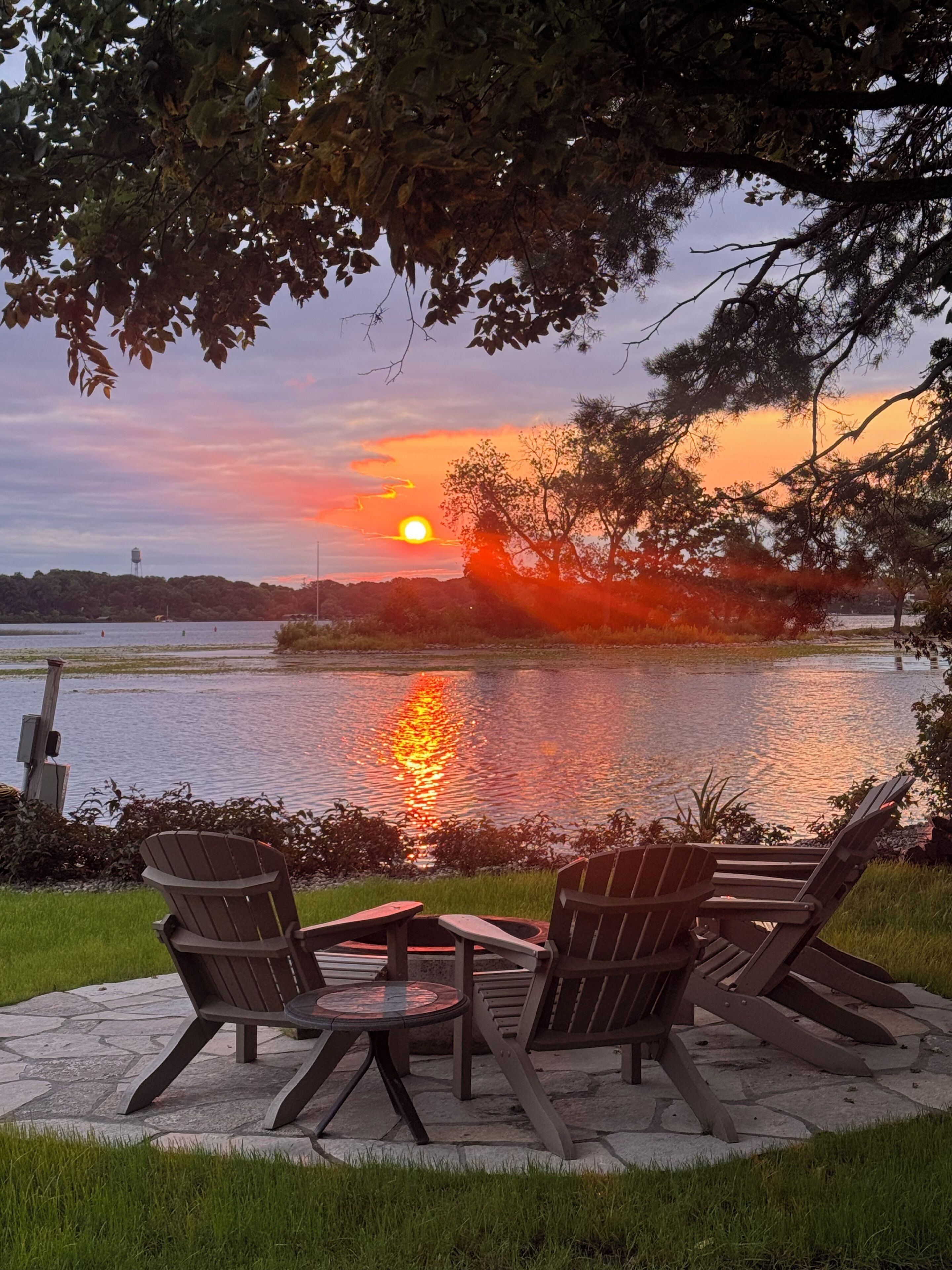 Vista al atardecer sobre un cuerpo de agua, con sillas y una mesa en un patio de piedra.