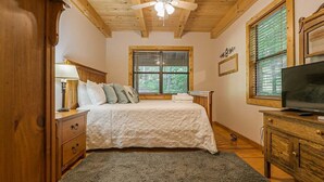 A bedroom with a bed, wooden headboard, a ceiling fan, a TV, a window with blinds, and a wooden dresser.