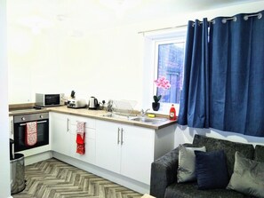 A compact kitchen with white cabinets, a microwave, a toaster, and a sink. There's a window with blue curtains and a potted plant on the counter. - Sigma Central Flat 5 a short walk away from Ferry Terminal and local amenities.. (North Shields)