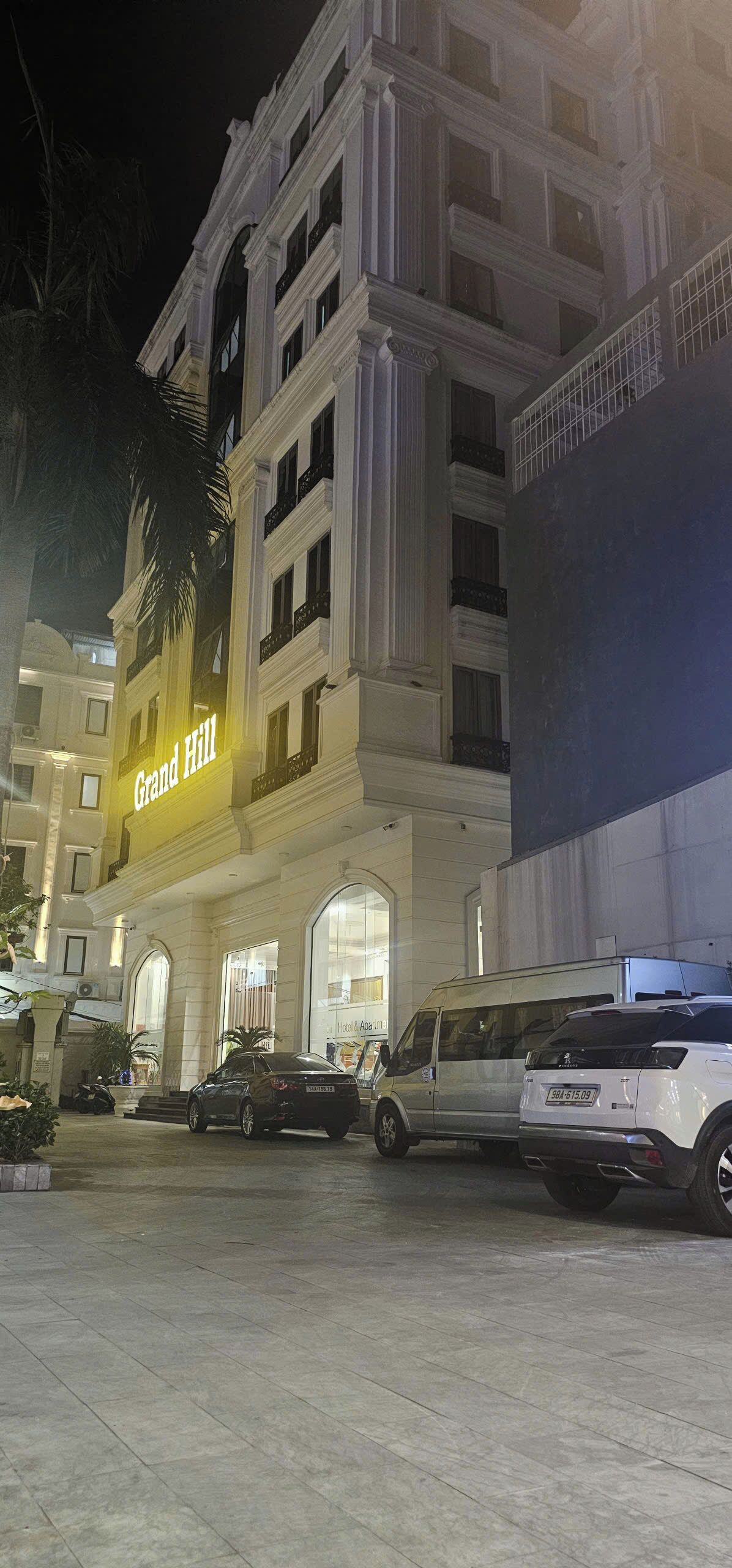 A hotel with a prominent 'Grand Hill' sign, multiple balconies, and a parking area with several cars.