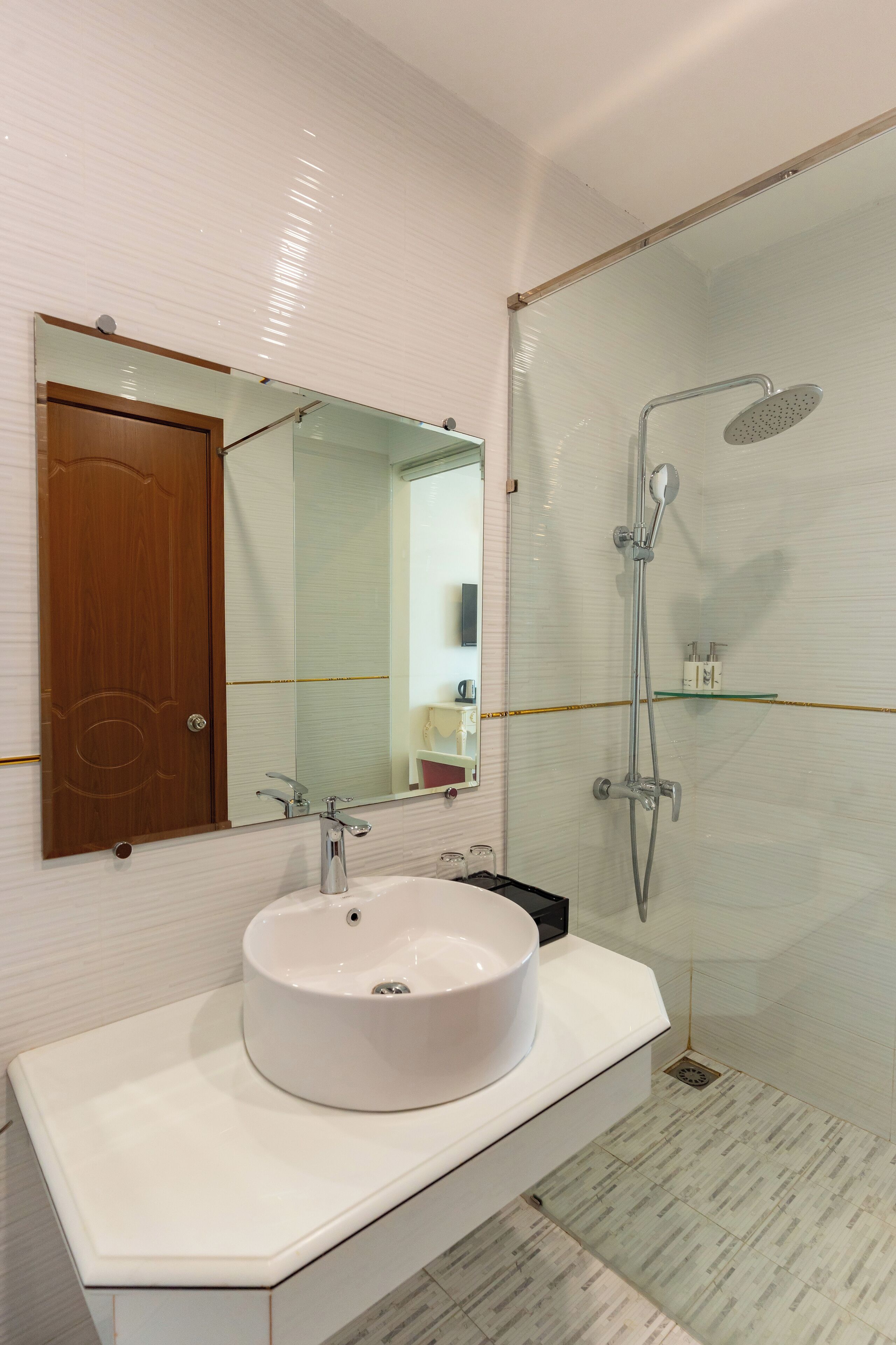 Un baño con lavamanos blanco, puerta de madera, espejo grande y cabina de ducha con paredes de vidrio.