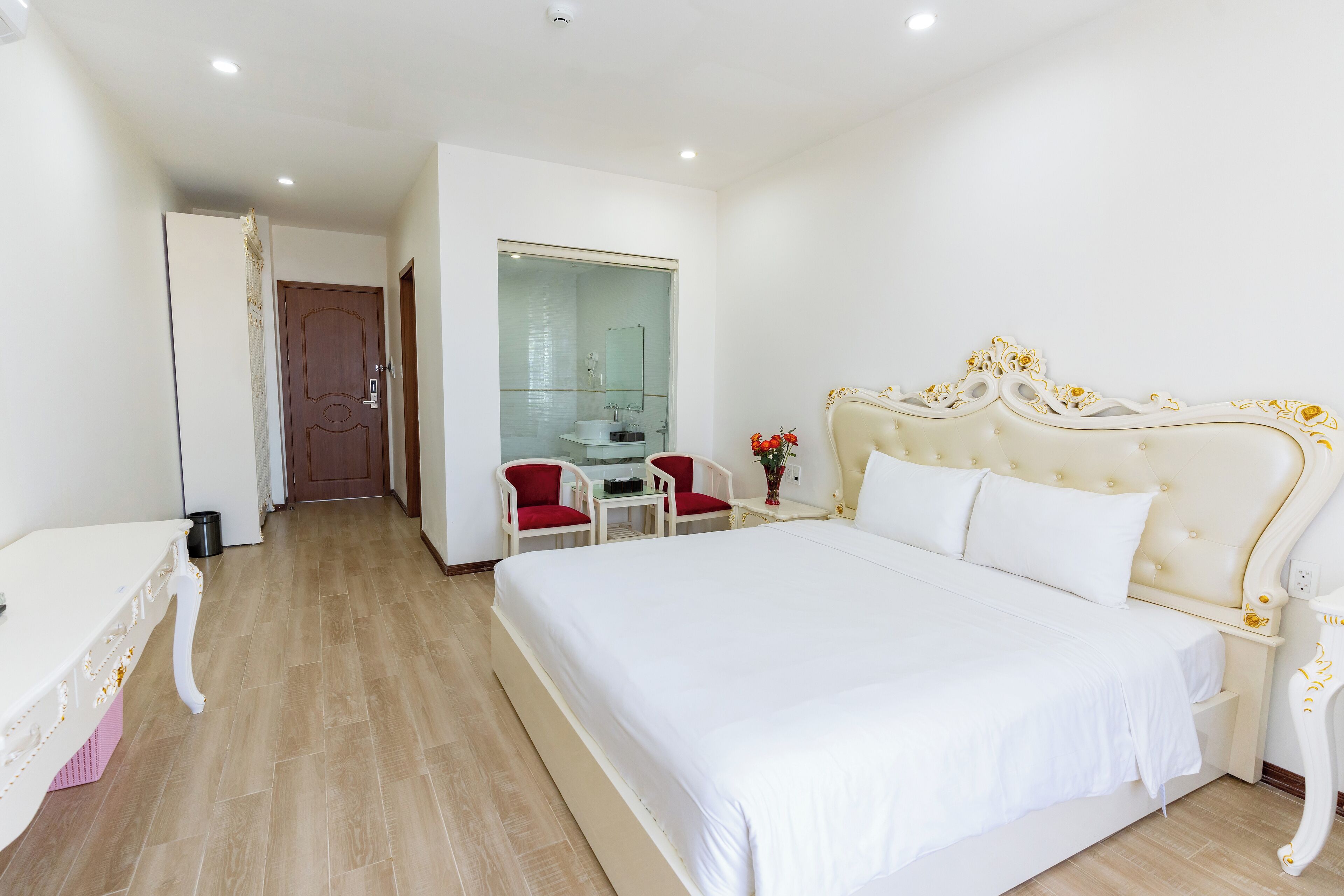 A hotel room with a large bed, a desk with two chairs, a bathroom with a glass door, and a wooden door.