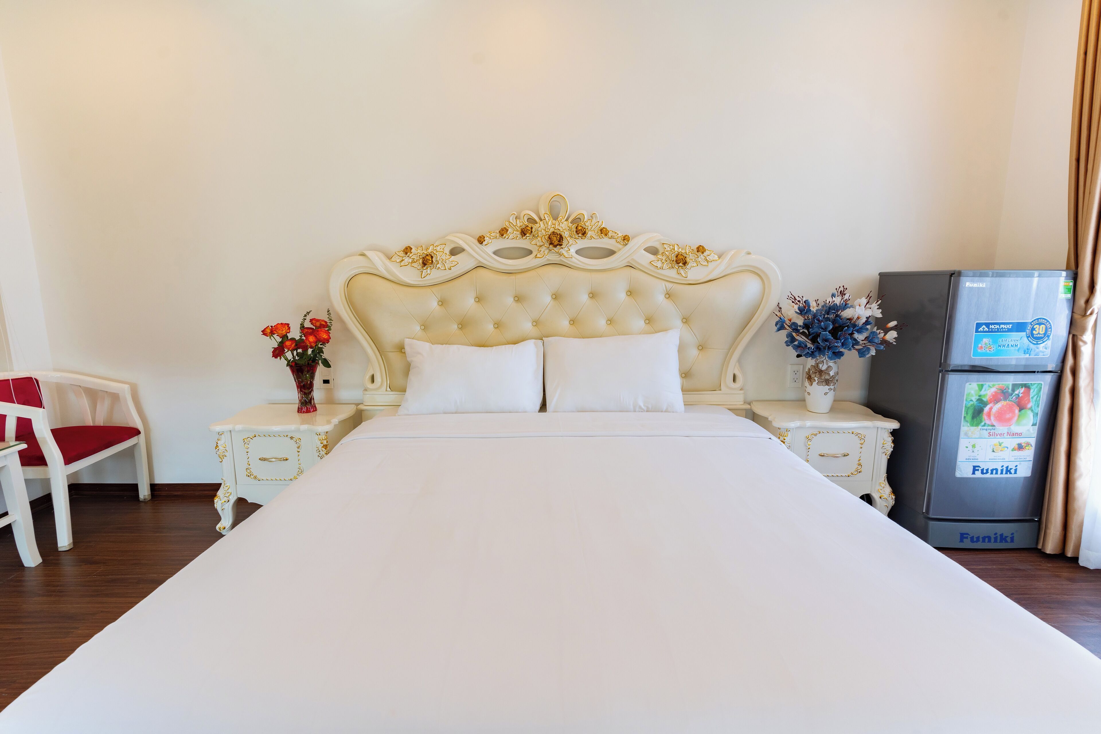 Una cama bien tendida con un cabecero capitonado, almohadas blancas y una colcha blanca. A la derecha, una mesita de noche con un florero y un refrigerador.