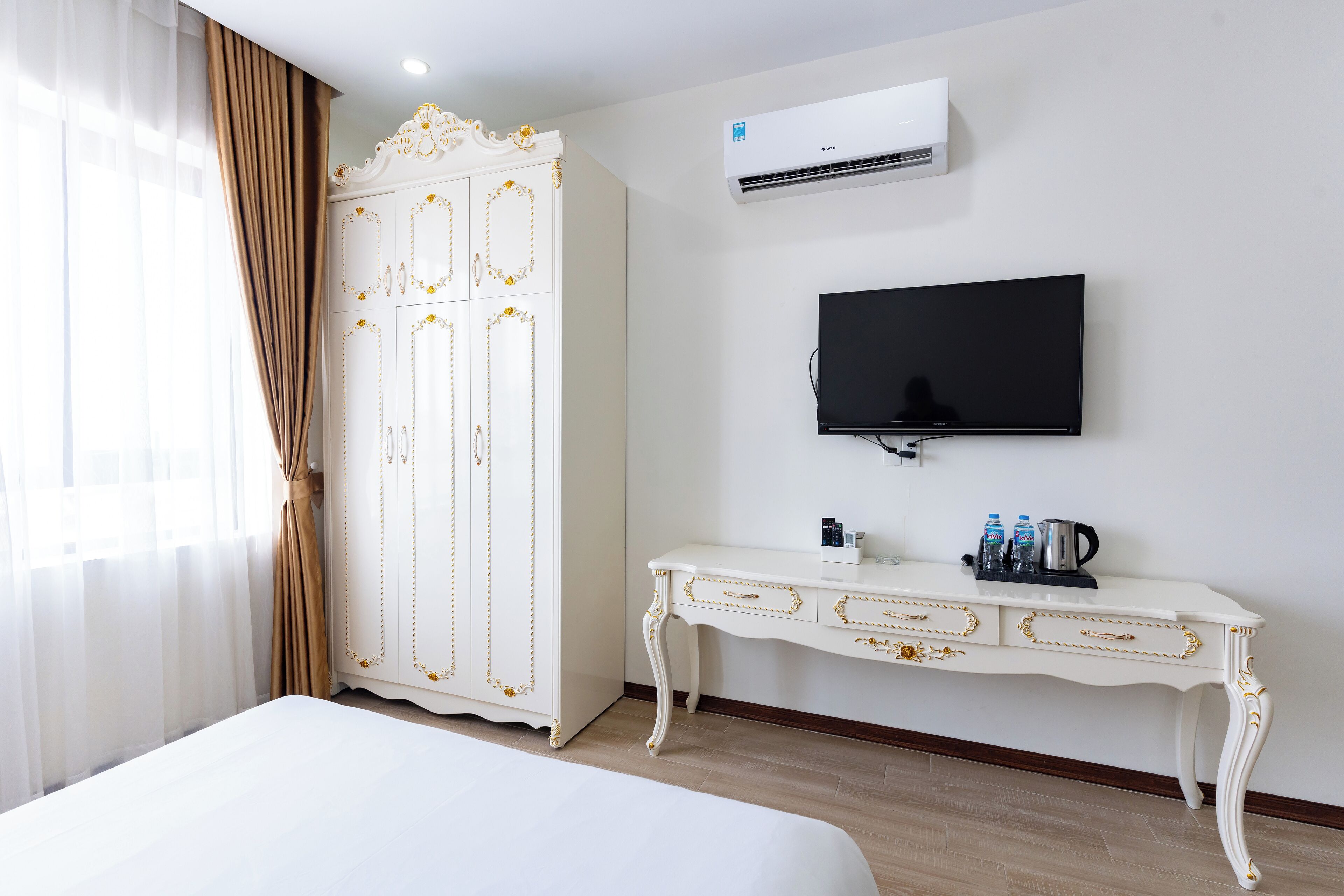 Habitación de hotel con cama, armario blanco con detalles dorados, televisor de pantalla plana, mesita con cafetera y botellas, y unidad de aire acondicionado en la pared.