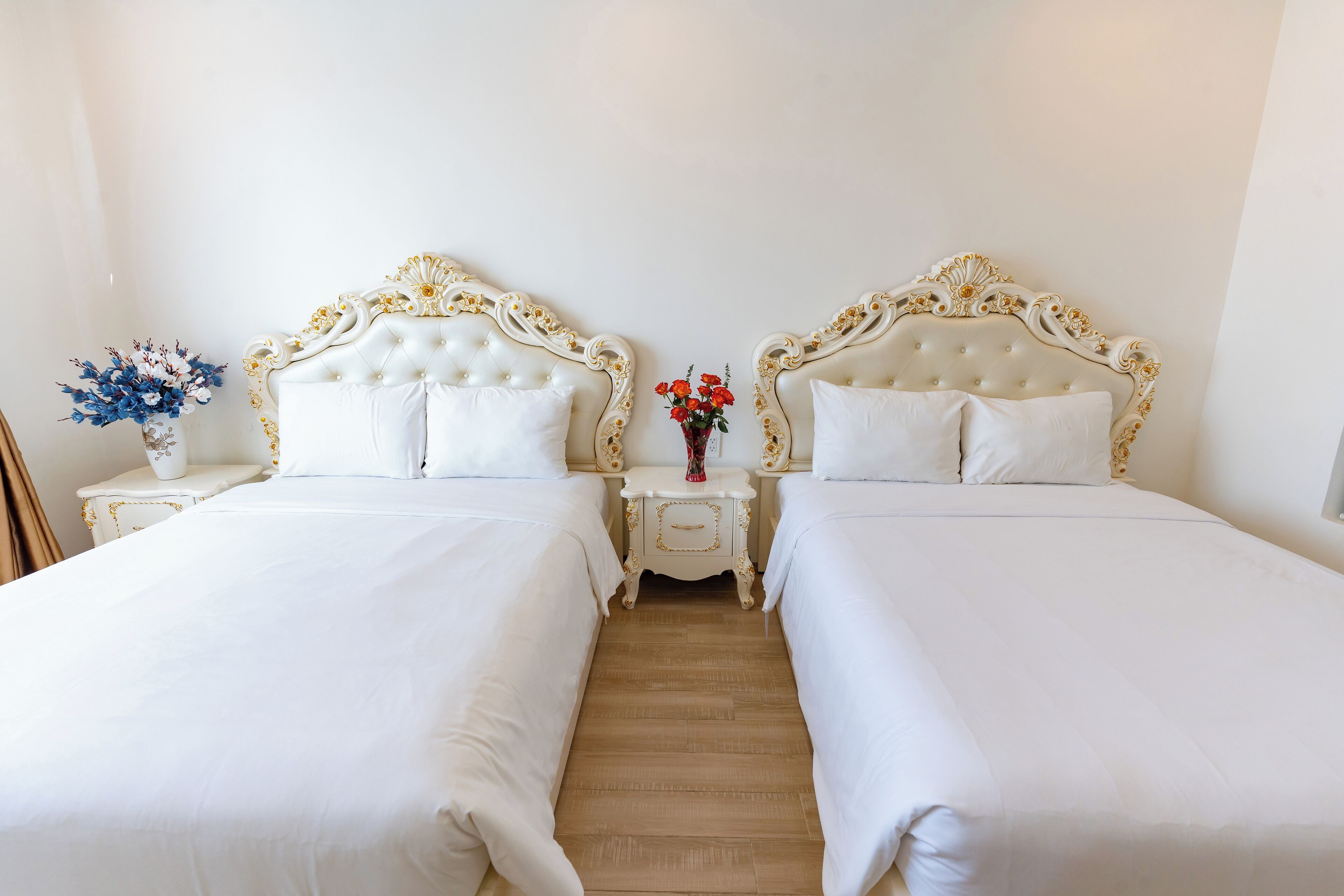 A double room with two beds, each with a white headboard and matching pillows. There is a nightstand with a vase of flowers between the beds.