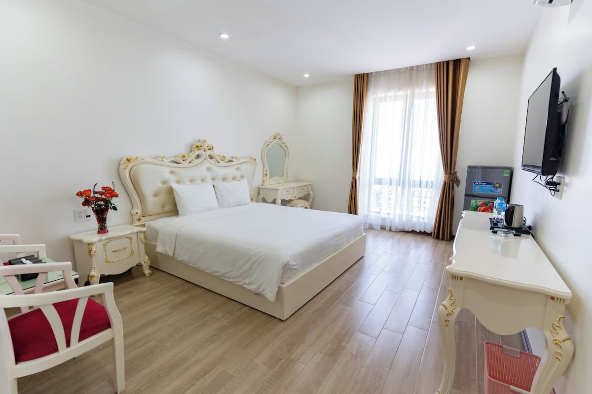 A hotel room with a large bed, a bedside table with a TV, a desk, a chair, and a vase with flowers.