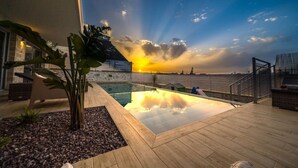 Ein Pool auf dem Dach mit freiem Blick auf den Sonnenuntergang über dem Meer, umgeben von Holzterrasse und einigen Pflanzen.
