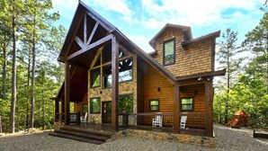 A log cabin with a stone foundation, large windows, and a porch with chairs. - New! Grand Hideaway| Secluded|6 Bedroom (Broken Bow)