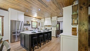 A modern kitchen with white cabinetry, a central island with bar stools, stainless steel appliances, and a wooden ceiling. - New! Grand Hideaway| Secluded|6 Bedroom (Broken Bow)