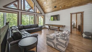 A spacious living room with a large leather sofa, a patterned ottoman, and a wooden coffee table. There is a flat-screen TV mounted on the wall and a wooden bench. Large windows provide natural light and views of the surrounding forest. - New! Grand Hideaway| Secluded|6 Bedroom (Broken Bow)